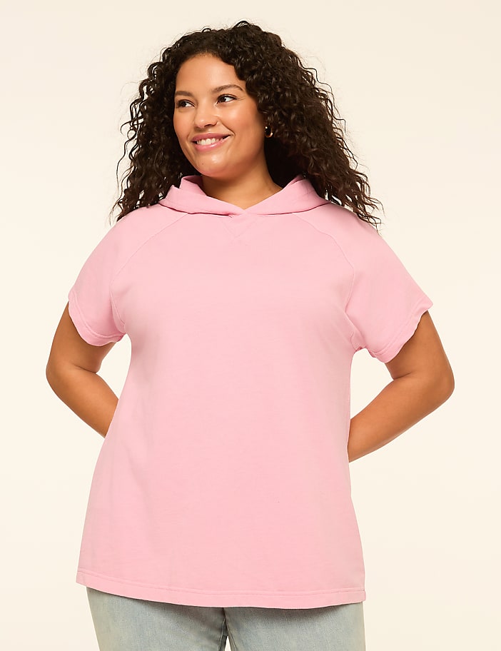 A smiling woman with curly dark hair stands in the foreground wearing a pink short-sleeved hoodie against a plain, light-colored background.