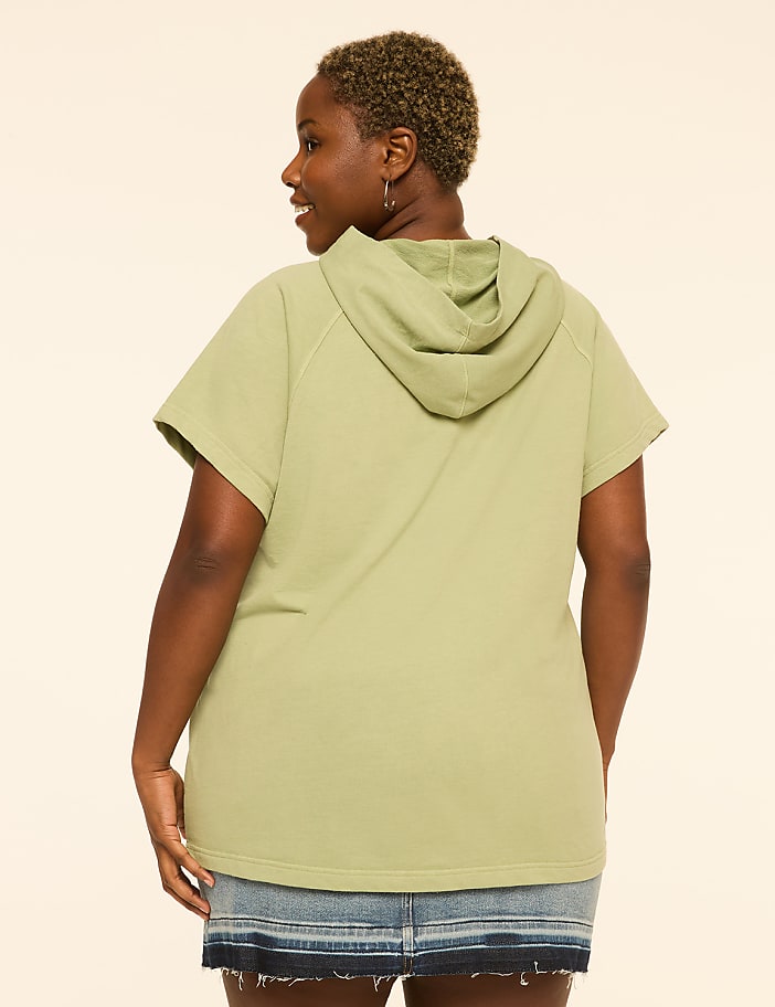 A woman in a light green short-sleeved hoodie and denim skirt stands against a plain, light-colored background.
