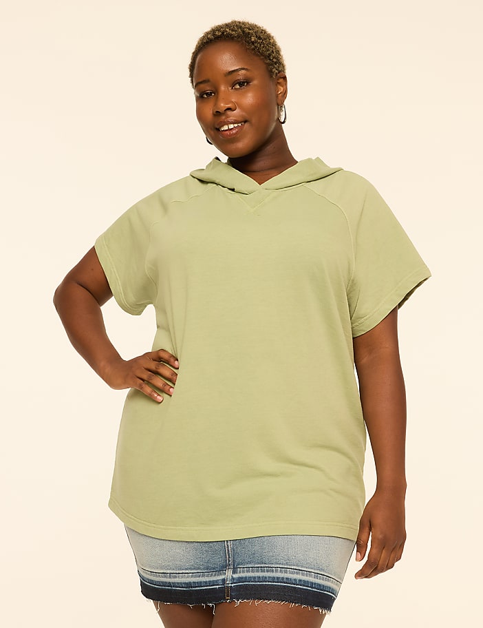A woman with short blonde hair and a light green short-sleeved hoodie stands with her hand on her hip against a plain, light-colored background.