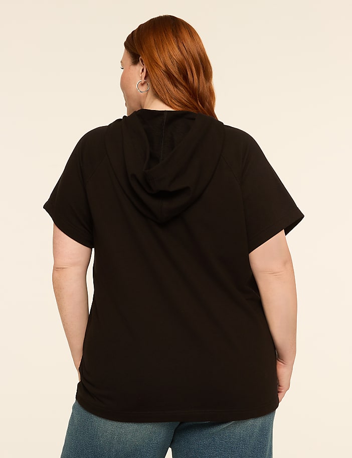 A woman with red hair wearing a black hooded t-shirt and blue jeans stands against a plain, light-colored background.