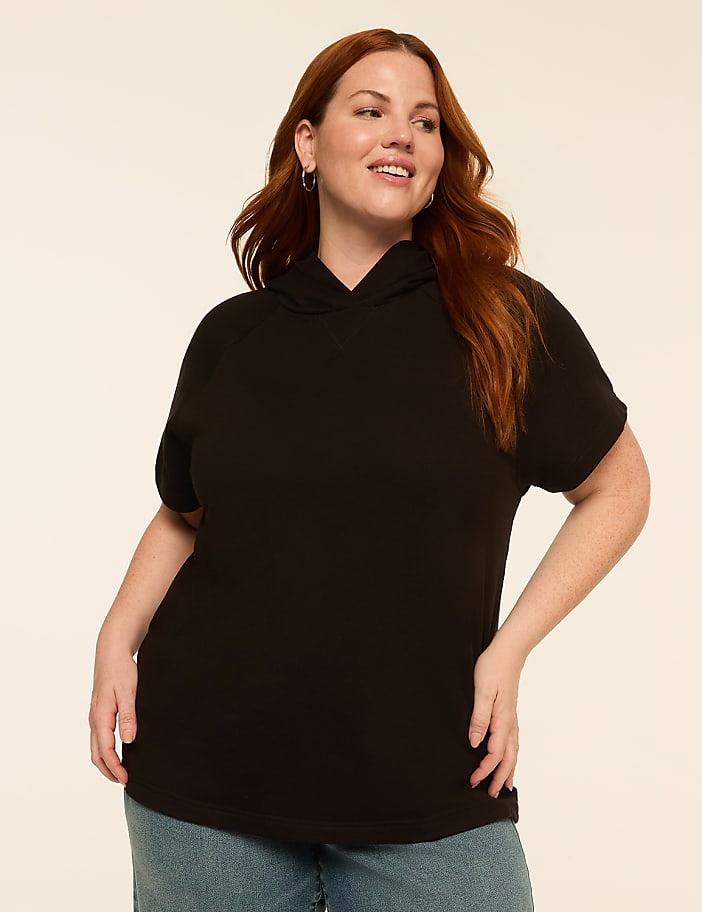 A smiling woman with red hair in a black short-sleeved hoodie and blue jeans stands against a plain, light-colored background.