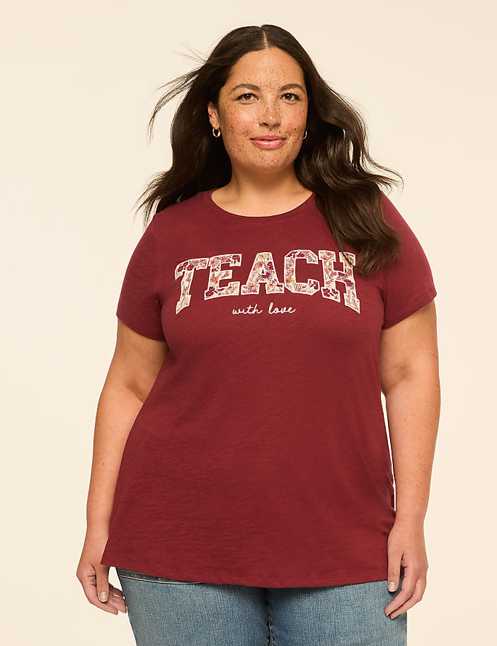 A woman with dark hair and freckles stands in the foreground wearing a maroon t-shirt with "TEACH with love" printed on it, against a plain, light-colored background.