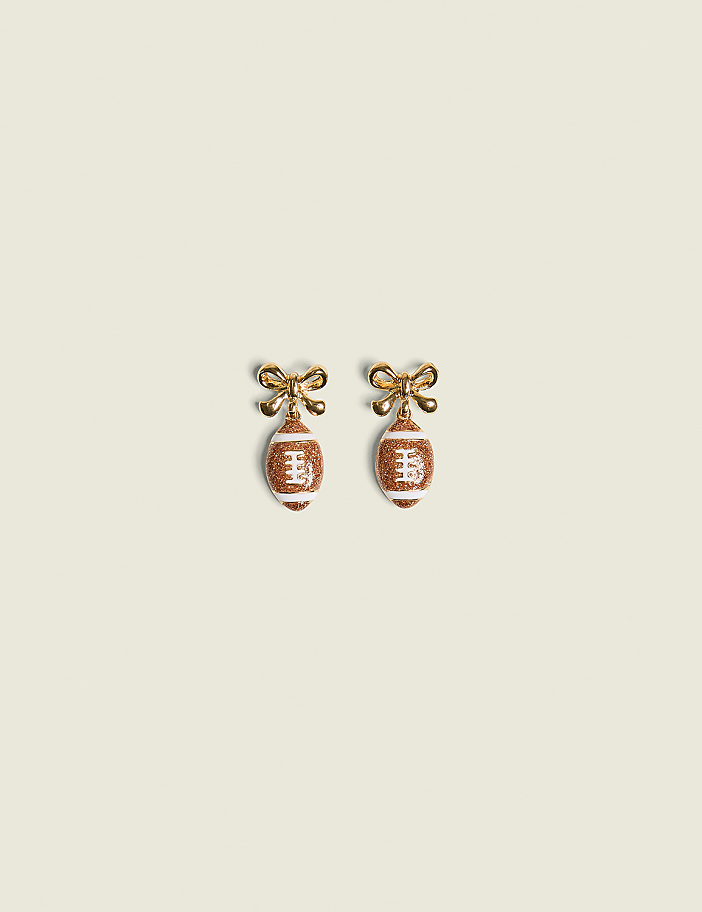 A pair of football-shaped earrings with gold bows hang against a plain, light-colored background.