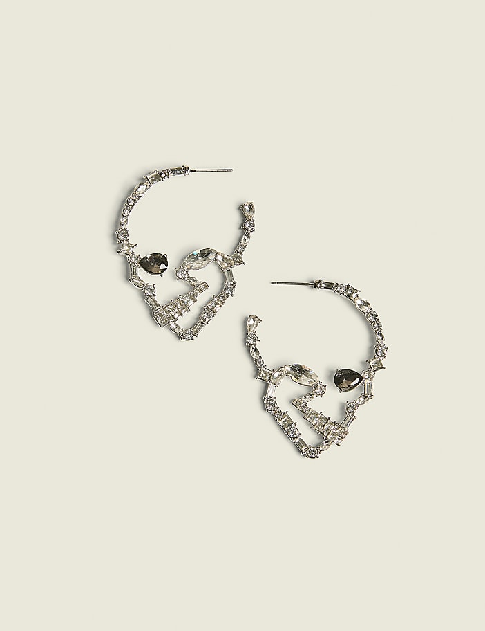 A pair of silver hoop earrings shaped like skulls with dark gem eyes rest on a pale yellow background.