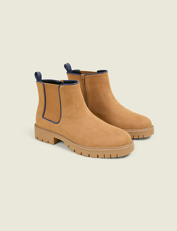 A pair of tan suede Chelsea boots with dark blue trim and thick lug soles are displayed against a plain, light gray background.
