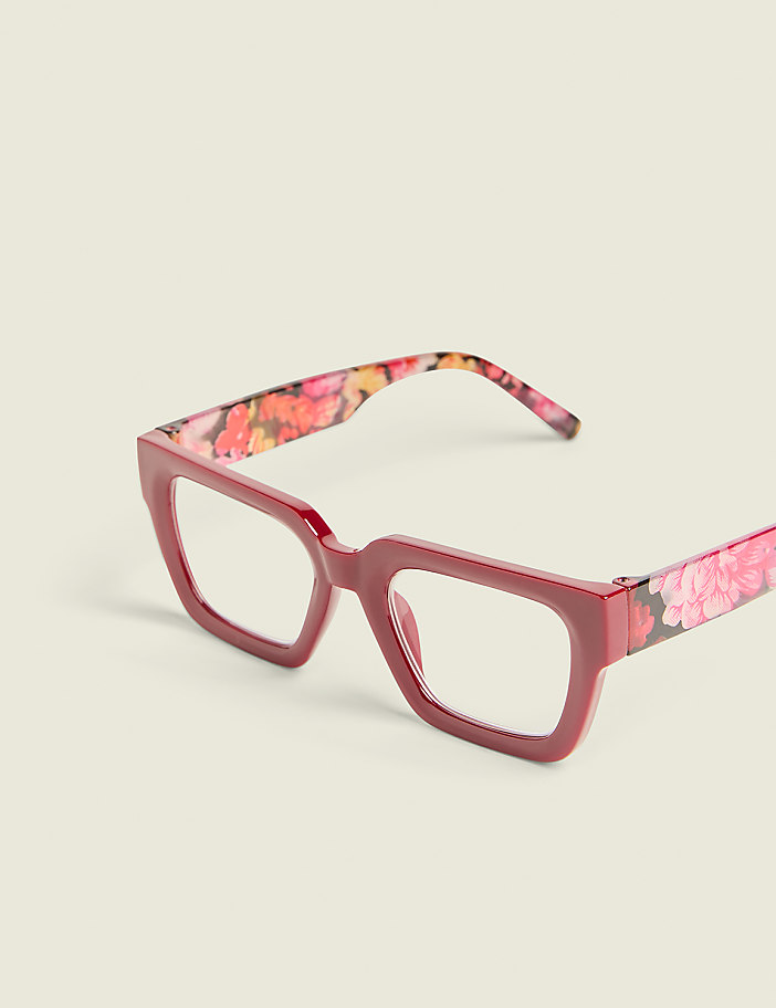 A pair of square-framed, dark pink glasses with floral patterned arms rests on a plain, light-colored background.