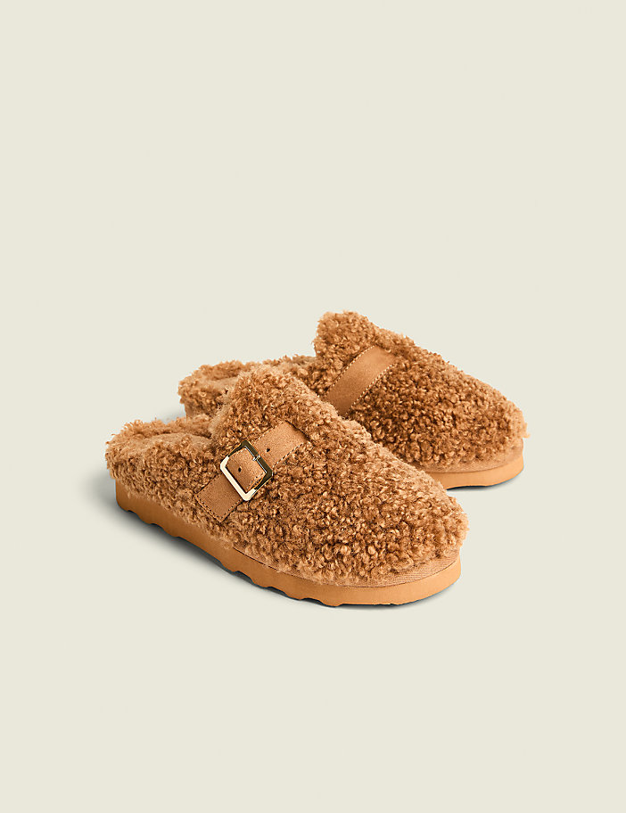 A pair of fuzzy brown clogs with buckle straps rests on a light beige background.