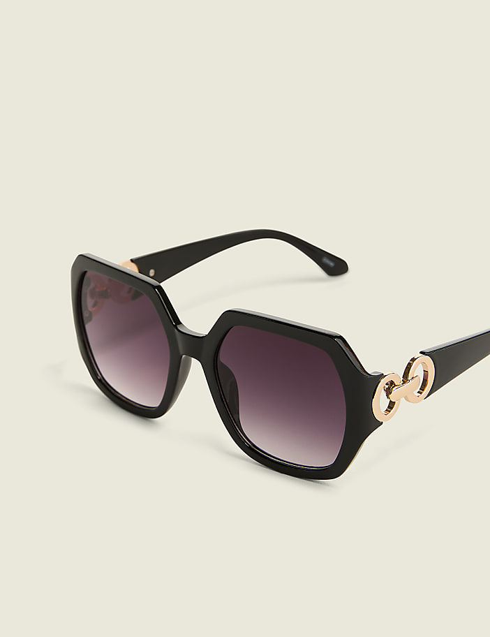 Black hexagonal sunglasses with gold chain accents rest on a plain, light-colored background.