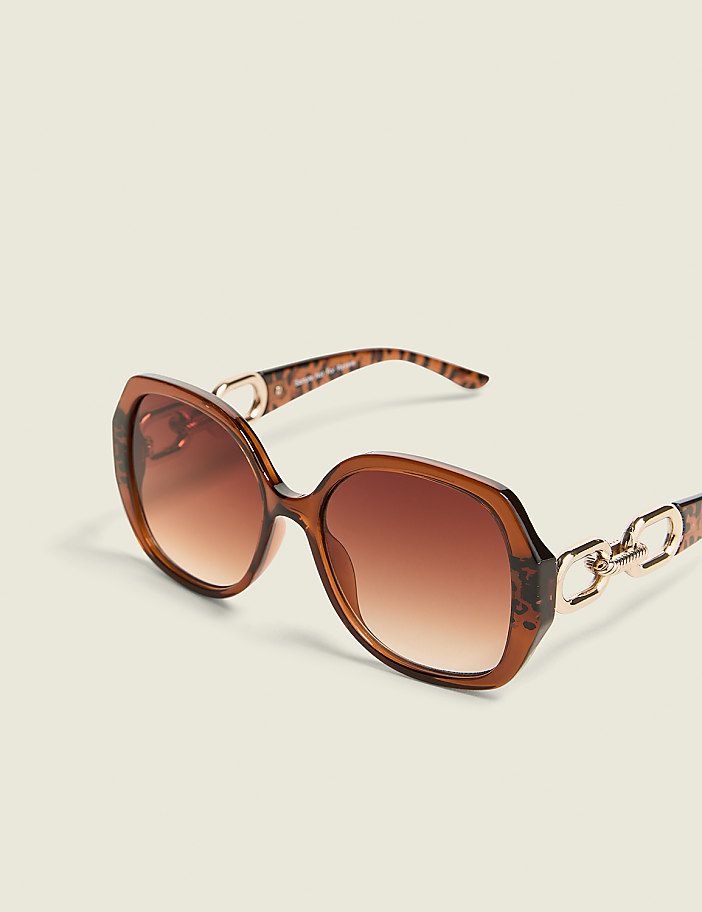 Oversized brown sunglasses with gold chain accents rest on a plain, light-colored background.