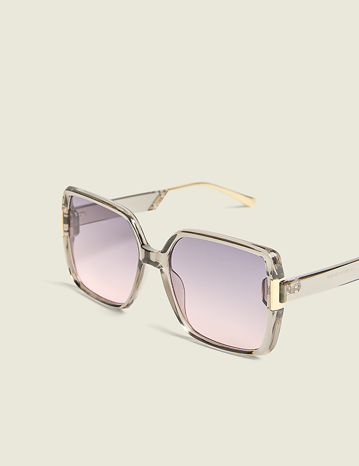 Oversized square sunglasses with a translucent grey frame and gradient pink lenses rest against a plain, light-colored background.
