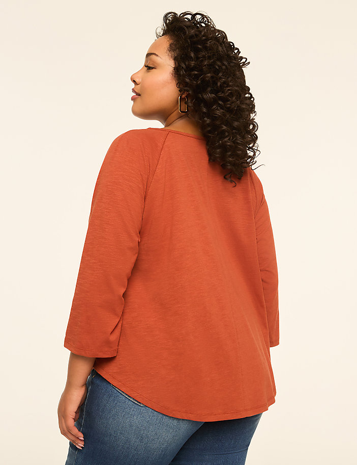 A woman with curly dark hair, wearing an orange shirt and blue jeans, stands against a plain cream background.