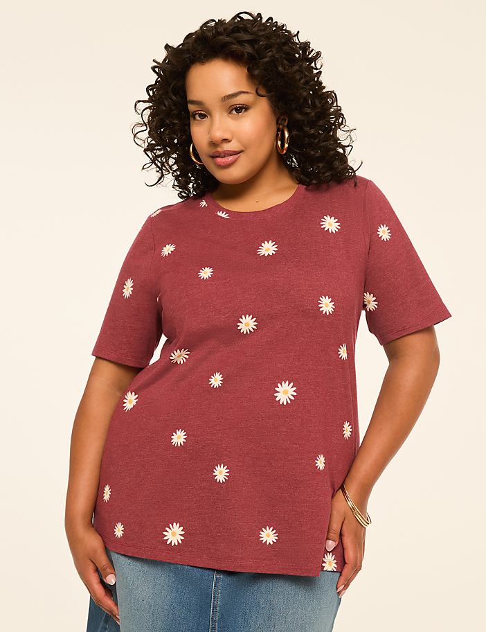 A woman with curly dark hair and hoop earrings poses in a maroon t-shirt with a daisy print against a plain, light-colored background.
