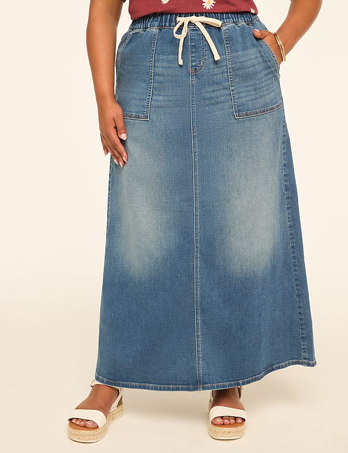 A woman in a denim maxi skirt and white sandals stands against a plain, light-colored background.