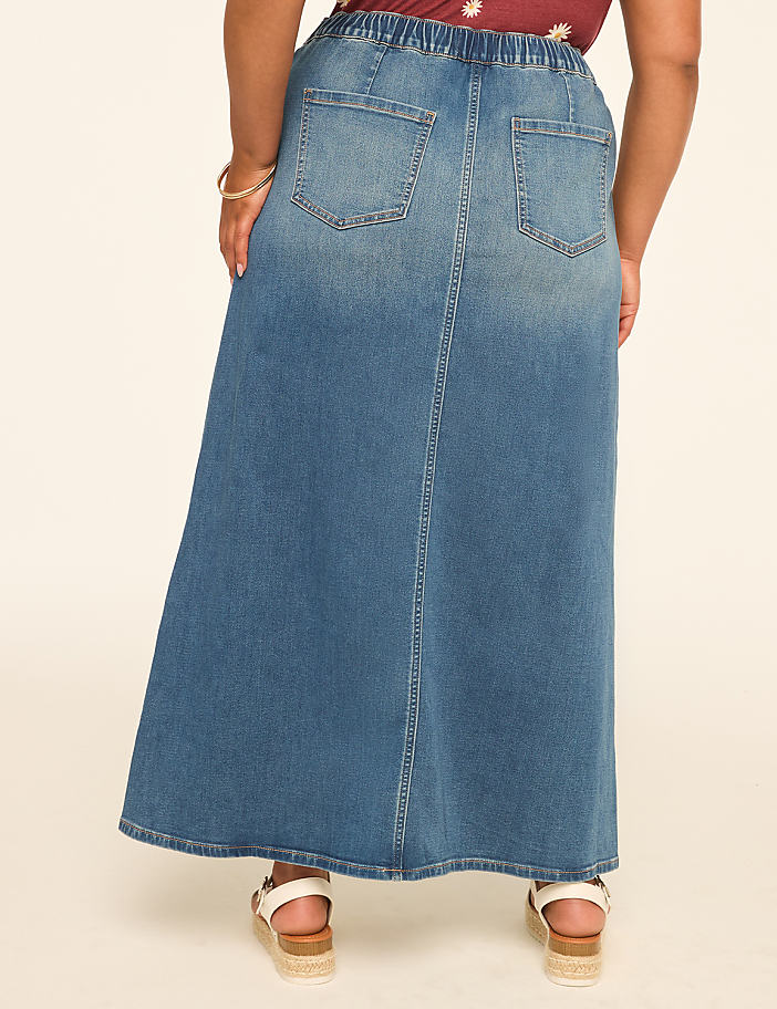 A person wearing a long denim skirt stands against a plain, light-colored background.