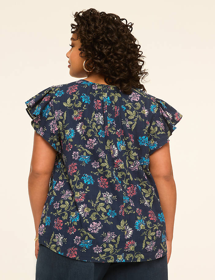 A woman with curly dark hair stands facing away from the camera, wearing a dark blue floral print top with ruffled sleeves, against a plain, light beige background.