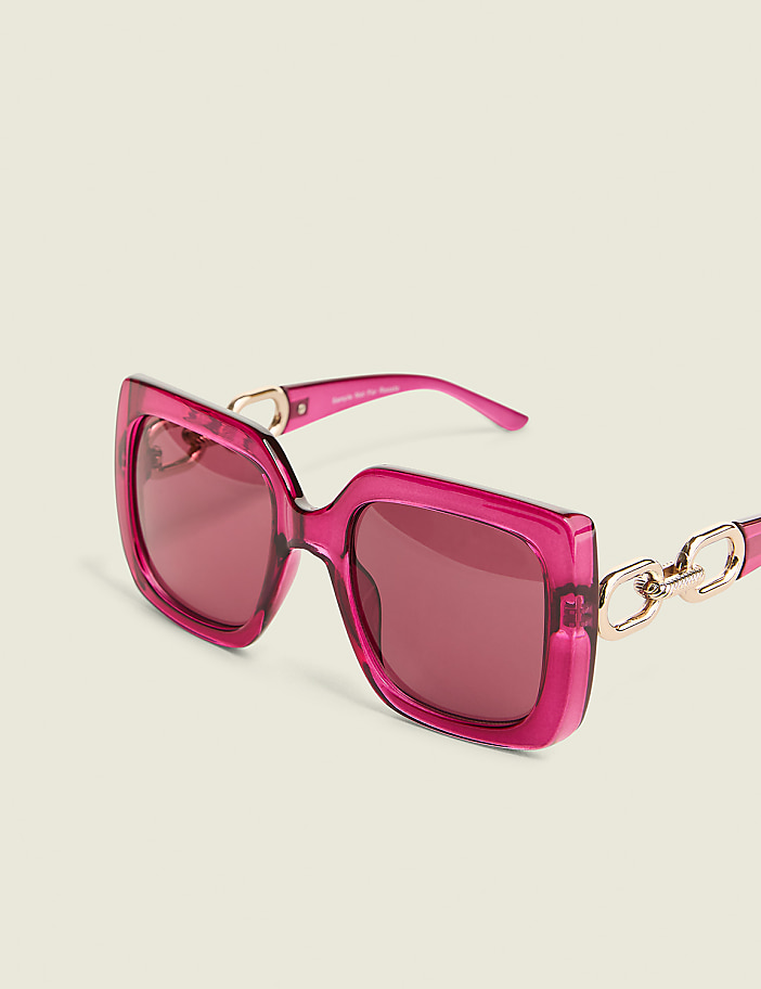 A pair of oversized, square, pink sunglasses with gold chain accents rests on a plain, light-colored background.