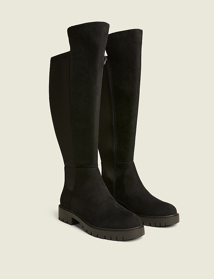 A pair of black suede knee-high boots with a zipper and a thick sole stand on a light brown background.