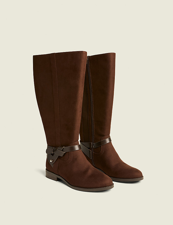 A pair of brown suede knee-high boots with decorative straps are displayed against a plain, light-colored background.