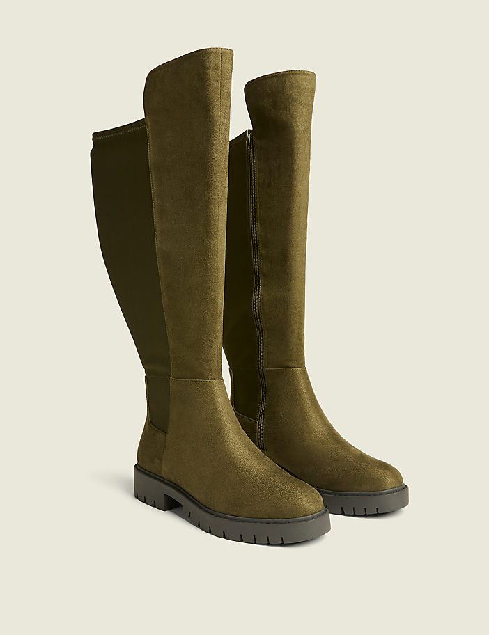 A pair of olive green, knee-high boots with a suede-like finish and a ribbed back panel stand on a plain, light beige background.