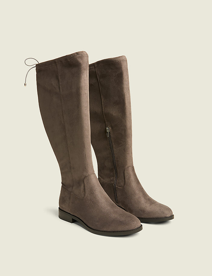 A pair of taupe suede knee-high boots with a tie detail at the back are displayed against a plain, light beige background.