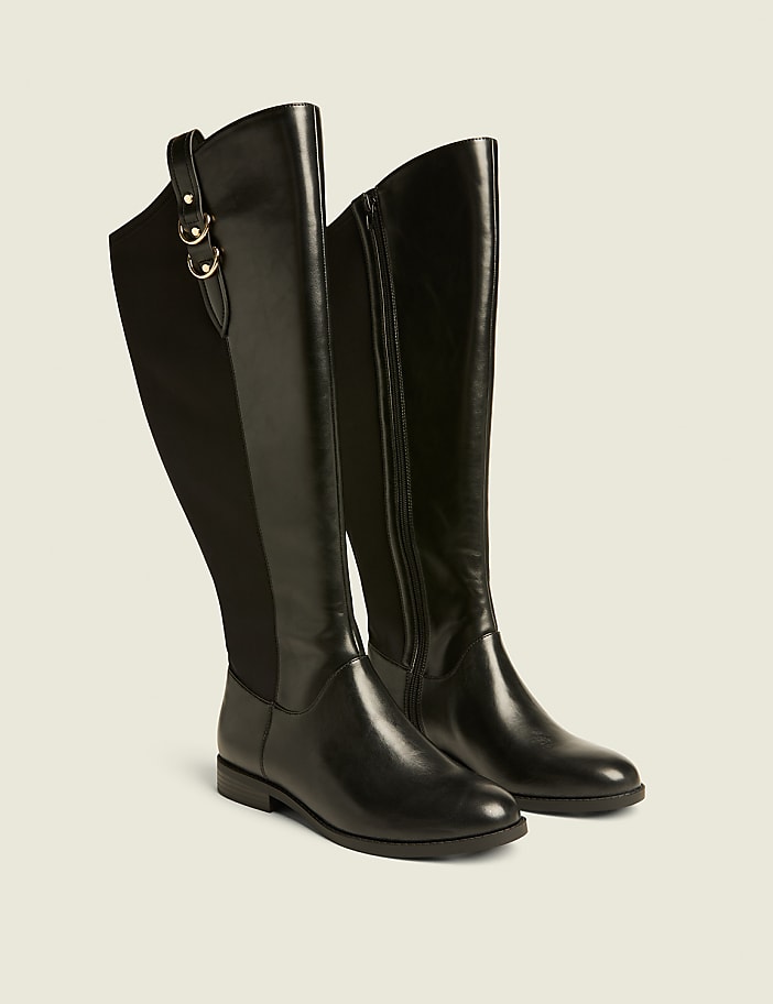 A pair of black leather knee-high boots with elastic side panels and gold buckle accents stand on a light beige background.