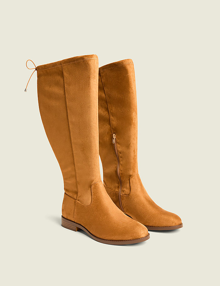 A pair of tan suede knee-high boots with a tie detail at the back are displayed against a plain, light brown background.