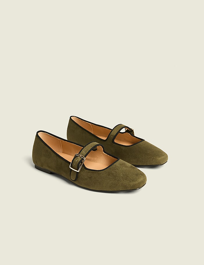 A pair of olive green suede Mary Jane flats with black trim and a gold buckle rests on a plain, light gray background.