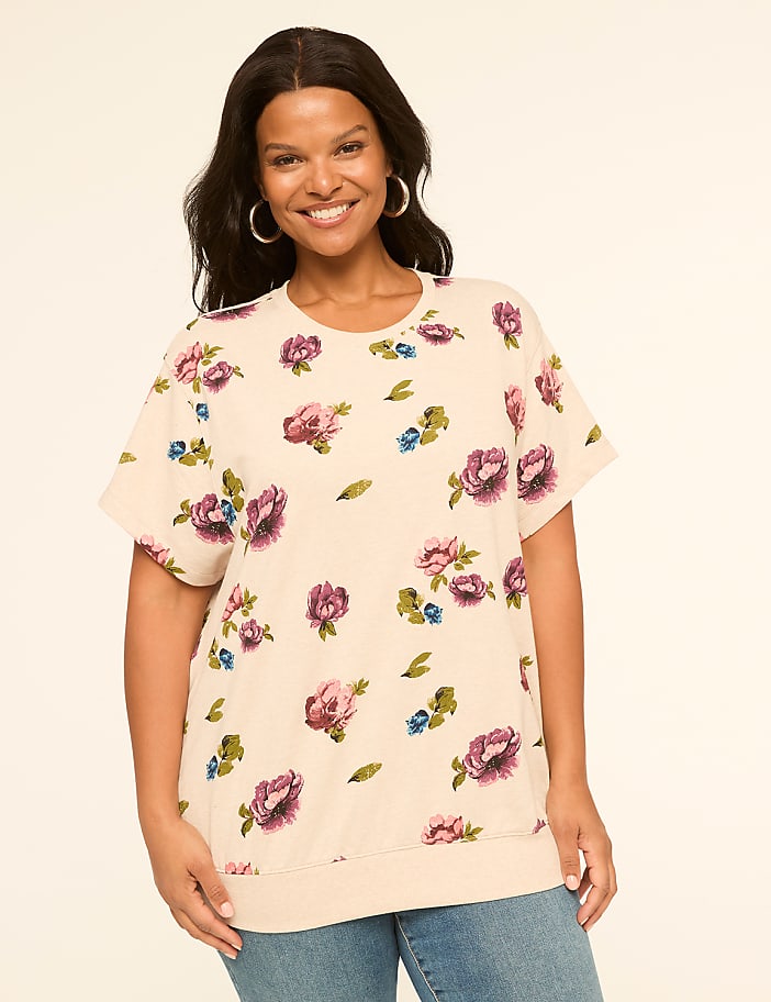 A smiling woman in a floral top stands against a plain, light-colored background.