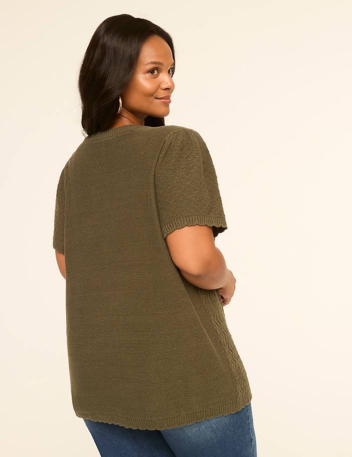 A woman in a textured olive green top and blue jeans stands against a plain, light-colored background.