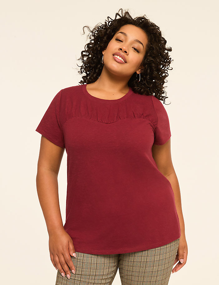 A woman with curly dark hair and a maroon t-shirt stands against a plain, light-colored background.