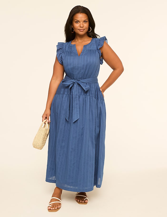 A woman in a blue striped dress stands against a plain, light-colored background, holding a straw bag.