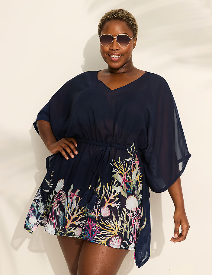 A woman in a navy blue cover-up with a coral print stands against a white background with palm leaf shadows.