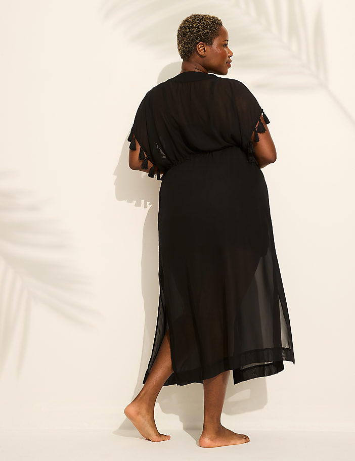 A woman in a black sheer dress stands with her back to the viewer, her bare feet on a white surface, while palm frond shadows are cast on the wall behind her.