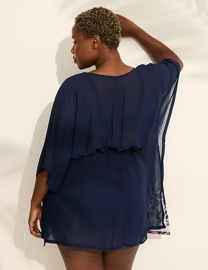 A woman in a navy blue sheer cover-up stands against a white wall with palm leaf shadows.