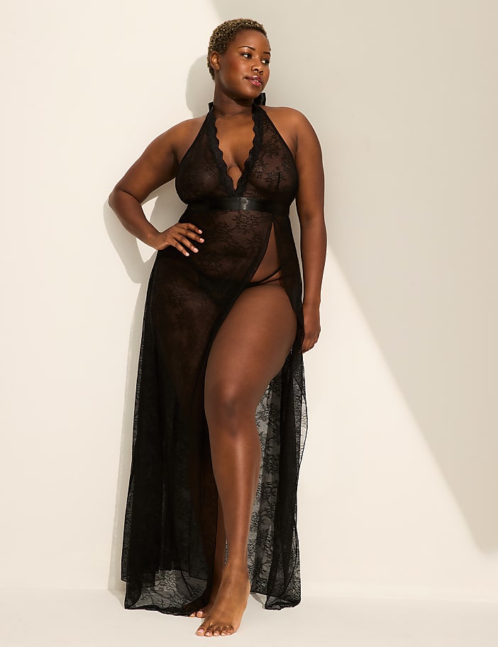 A woman in a sheer black lace gown stands against a plain, light-colored wall with a shadow cast on it.