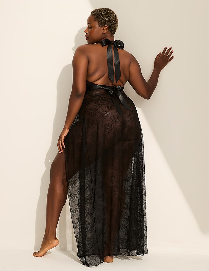 A woman in a sheer black negligee stands with her back to the viewer, leaning against a plain, cream-colored wall.