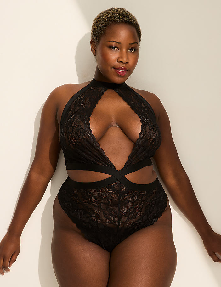A woman in a black lace bodysuit poses against a plain white wall with soft lighting.