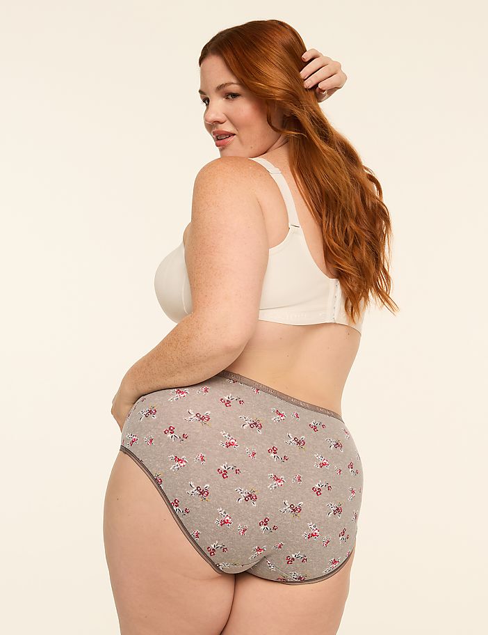 A woman with red hair in a white bra and floral underwear stands against a plain, light-colored background.