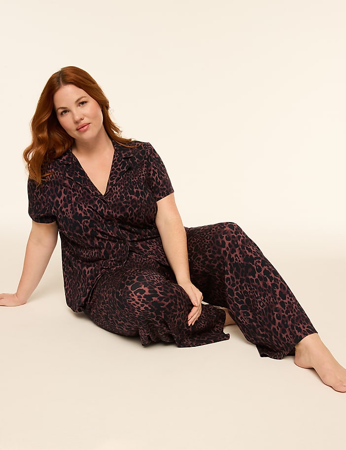 A woman with red hair sits on a light-colored floor wearing a leopard-print pajama set, with a plain, light background behind her.