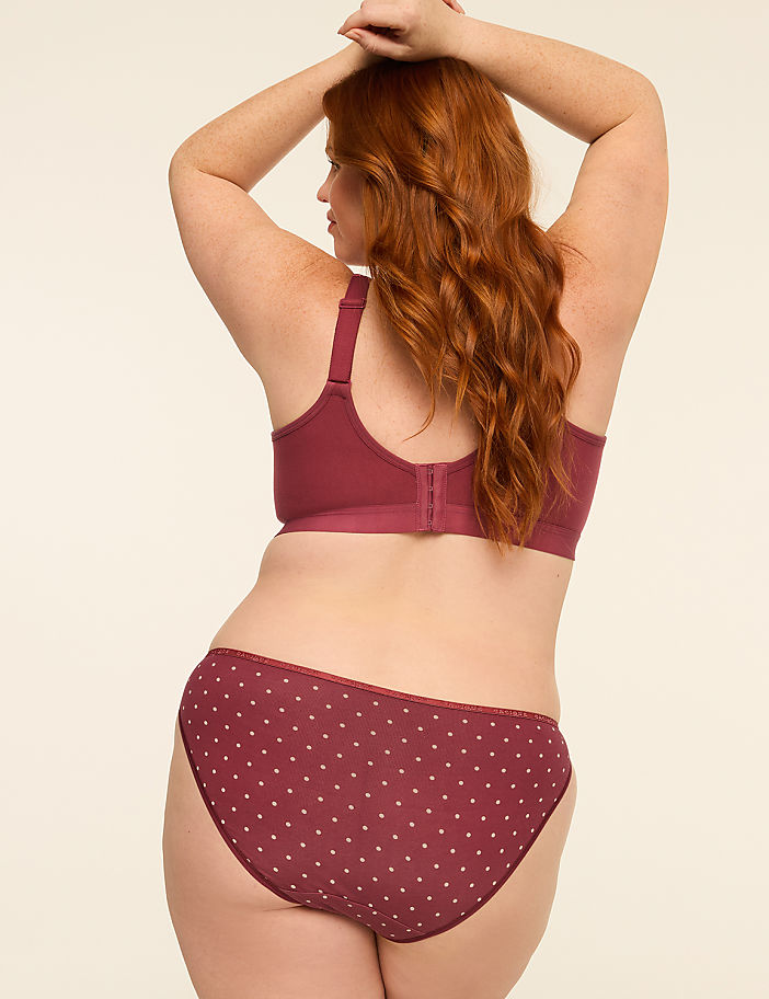 A woman with red hair stands with her back to the viewer, wearing a maroon polka dot bra and underwear against a plain, light-colored background.