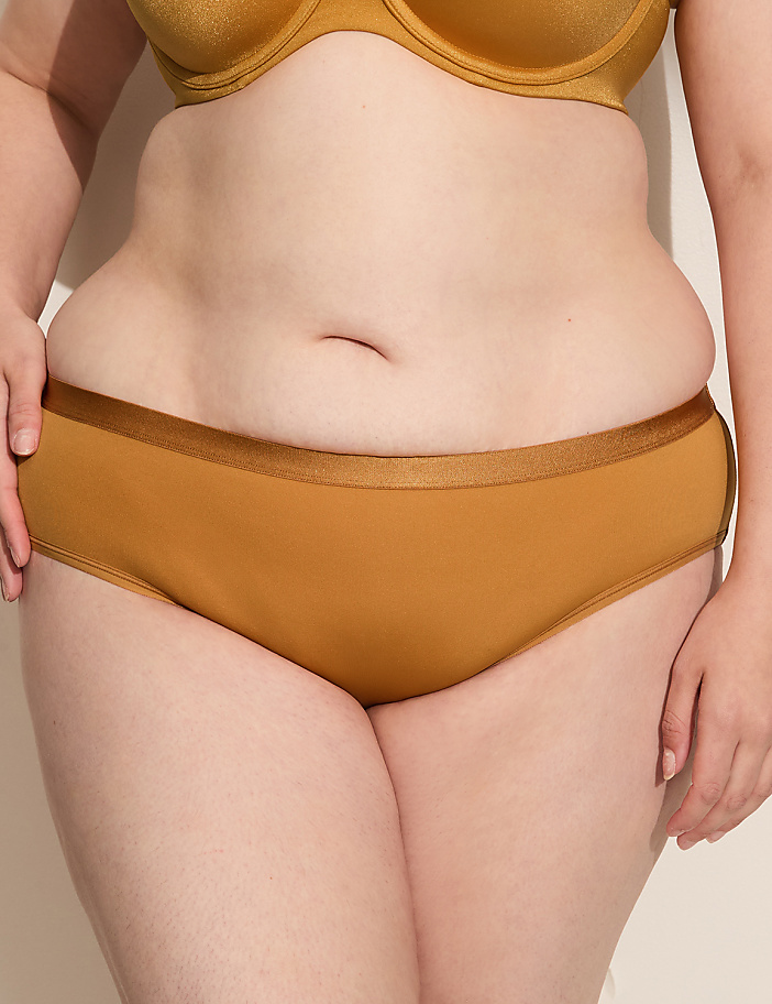 A person's midsection is shown in the foreground, wearing mustard-colored underwear and a matching bra, against a plain, light-colored background.