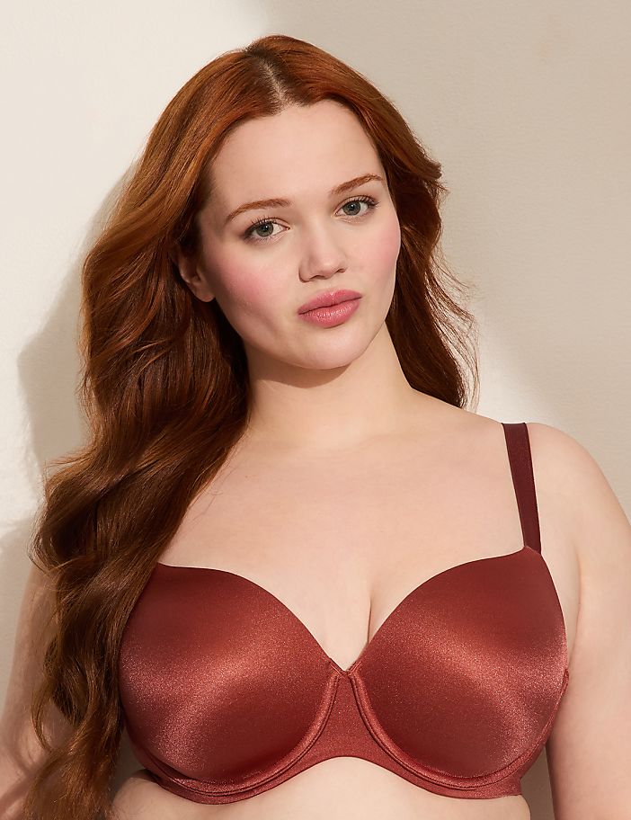 A woman with long, wavy red hair poses in a rust-colored bra against a plain, light-colored background.