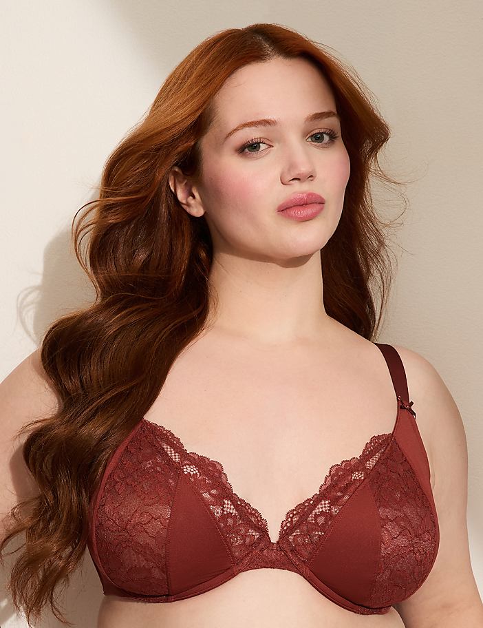 A woman with long, wavy red hair poses in a rust-colored lace bra against a plain, light-colored background.