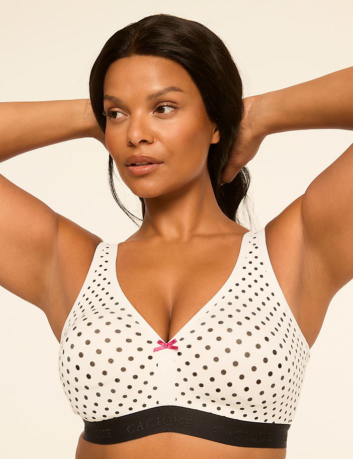 A woman with her hands behind her head wears a white polka dot bra against a plain, light background.