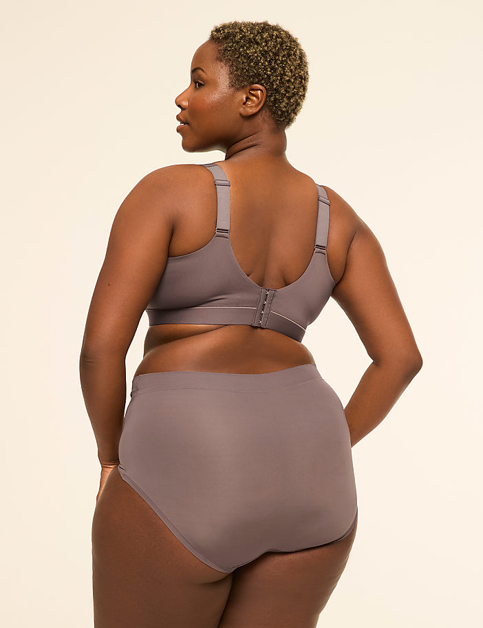 A woman with short, curly hair stands facing away from the camera in a taupe bra and matching high-waisted underwear against a plain, light-colored background.