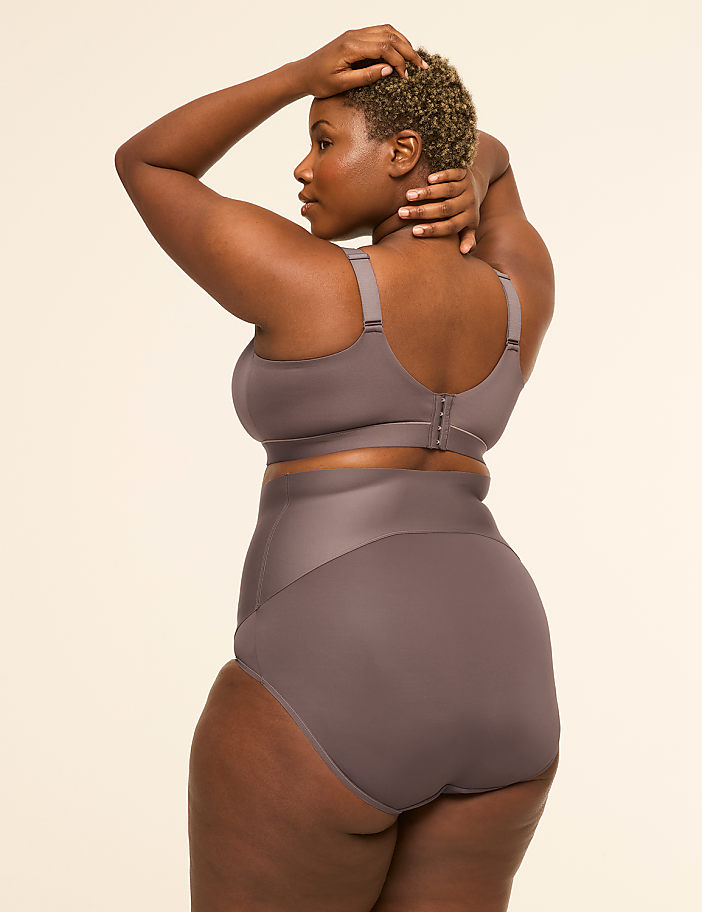 A woman in a grey bra and high-waisted underwear poses with her hands behind her head against a plain, light background.