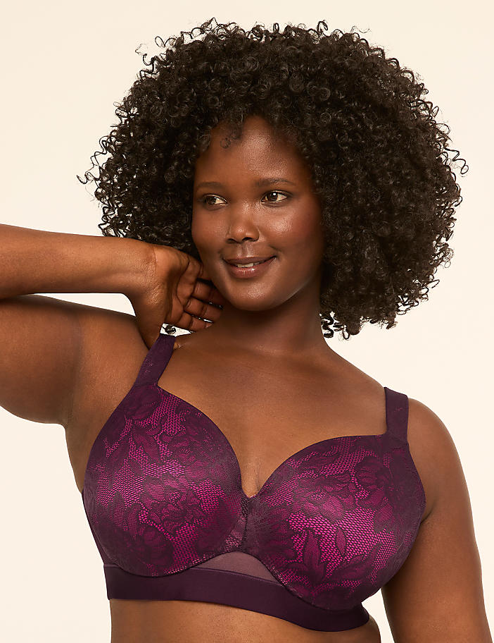 A woman with dark, curly hair smiles as she poses in a purple lace bra against a plain, light-colored background.