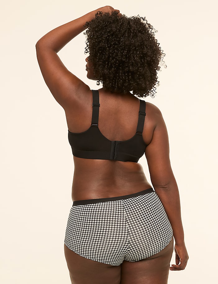A woman with dark curly hair, wearing a black sports bra and houndstooth patterned underwear, stands against a plain, light-colored background.