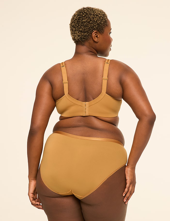 A woman in tan lingerie stands with her back to the viewer against a plain, light-colored background.