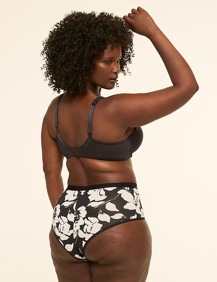 A woman with dark curly hair and dark skin, wearing a black bra and floral patterned underwear, stands against a plain, light-colored background.